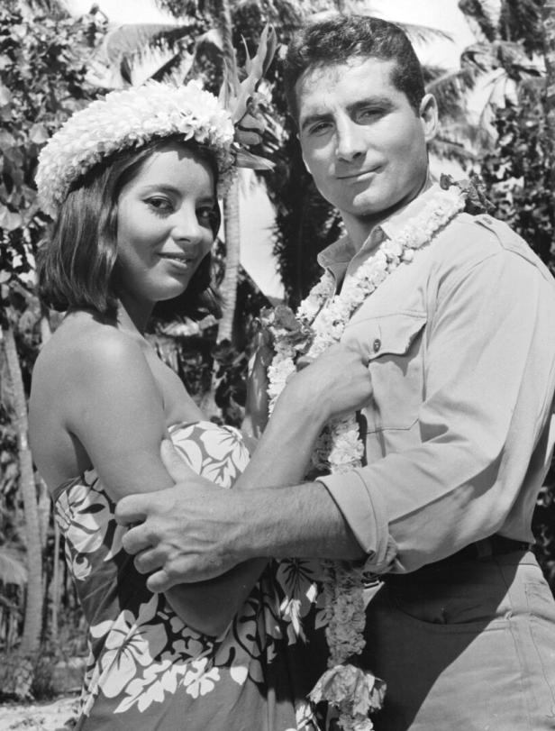 Ein Mann und eine Frau mit Blumenkränzen posieren vor tropischer Vegetation.