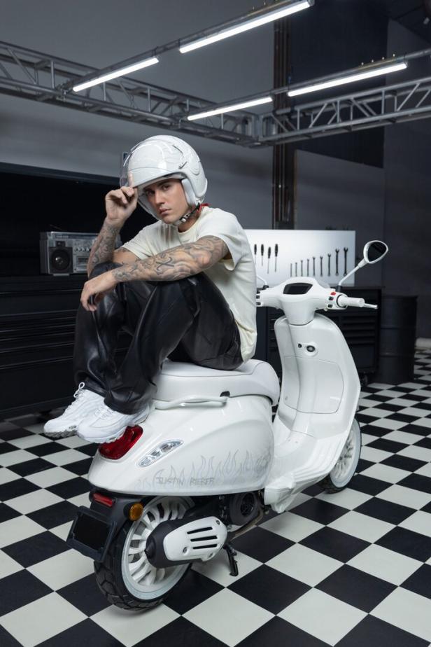 Justin Bieber sitzt mit Helm auf einer weißen Vespa in einer Werkstatt.