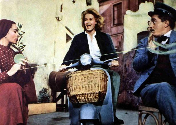 Eine junge Frau fährt auf einer Vespa, während zwei Personen mit einem Seil hantieren.