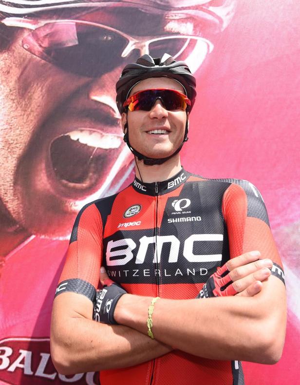 Ein lächelnder Radrennfahrer im Trikot von BMC Switzerland posiert vor einem Poster.