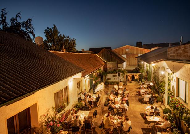 Auf dem Bild sieht man einen Innenhof eines Bauernhauses mit vielen Besucher:innen, die einen gemütlichen Abend genießen.