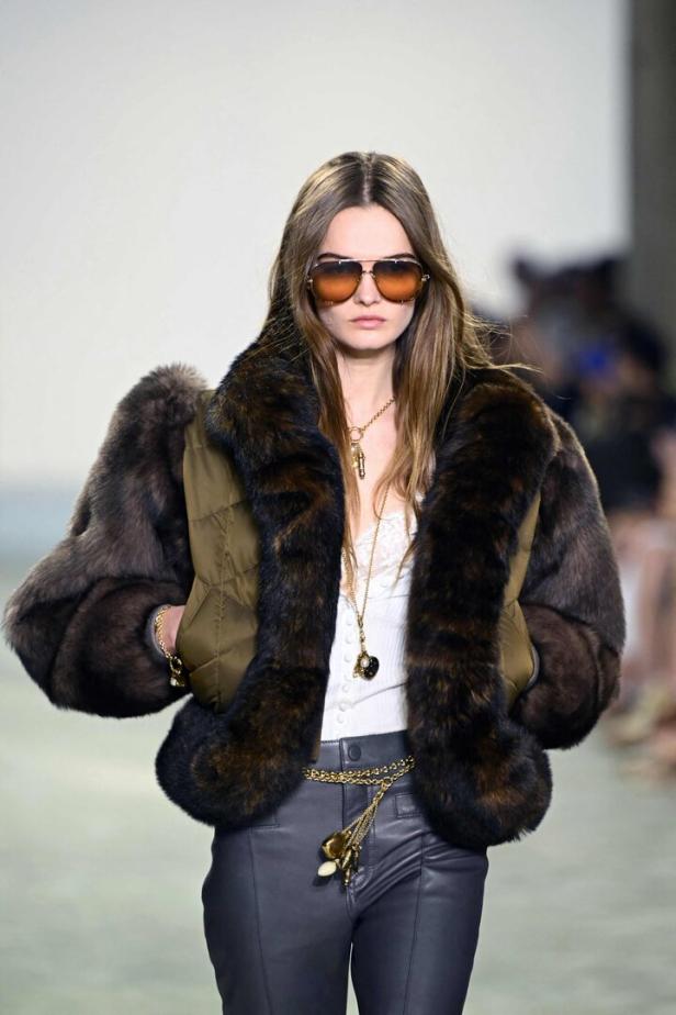 Ein Model präsentiert eine Jacke mit Pelzbesatz und eine goldene Kette auf dem Laufsteg.