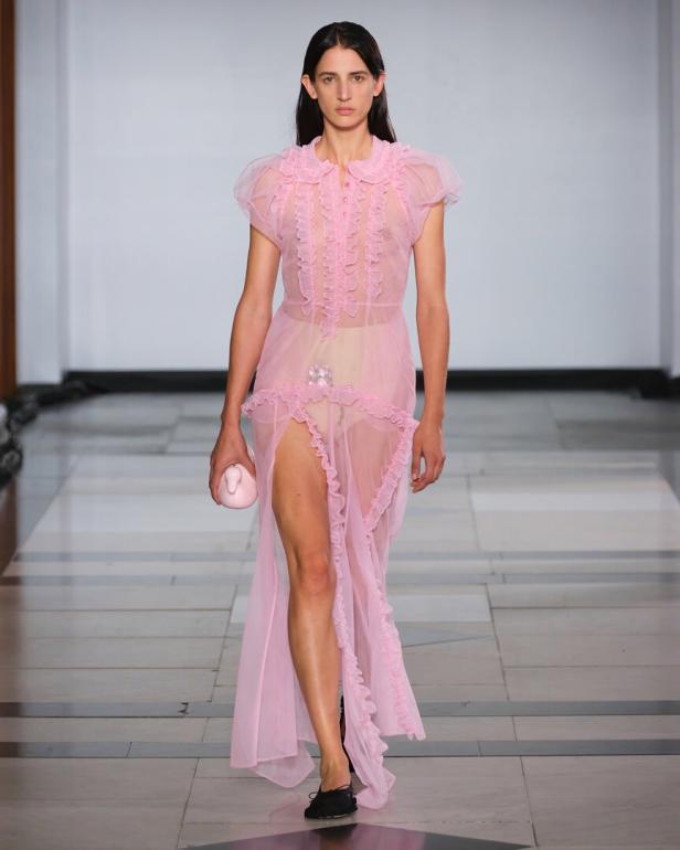 Eine Frau in einem transparenten, rosa Kleid mit Rüschen und einer kleinen, rosa Handtasche auf einem Laufsteg.