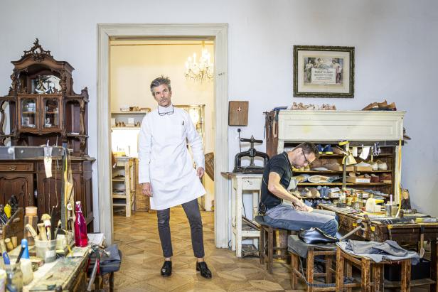 Ein Mann im weißen Kittel steht in der Werkstatt von Maßschuhe Scheer, während ein anderer an einem Schuh arbeitet.