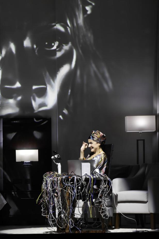 Eine Frau mit Turban sitzt an einem Tisch voller Kabel und arbeitet am Laptop. Im Hintergrund ist ein Gesicht projiziert.