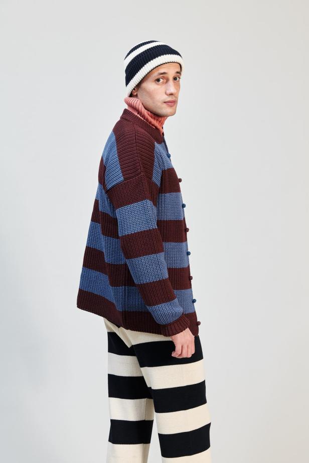Ein Mann mit geringeltem Cardigan, Hose und Mütze vor einem hellen Hintergrund.