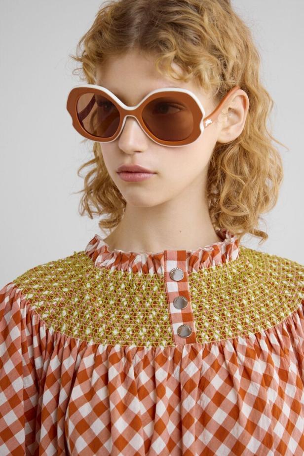 Eine junge Frau mit lockigem Haar trägt eine karierte Bluse und braune Sonnenbrille.