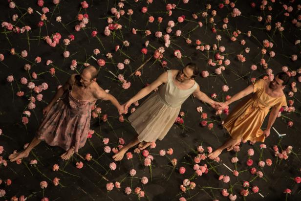 Drei Männer in Kleidern halten sich auf einer Bühne voller Blumen an den Händen.