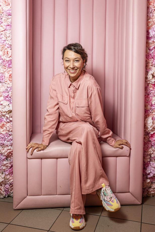 Ursula Strauss sitzt in einem rosa Sessel vor einer Wand aus Rosen.