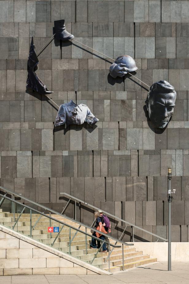 Skulptur an einer Fassade mit Uniformteilen und einem Kopf, darunter eine Treppe mit Passanten.