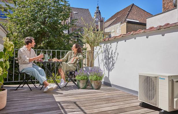 Zwei Personen sitzen auf einem Balkon, auf dem eine Klimaanlage zu sehen ist.
