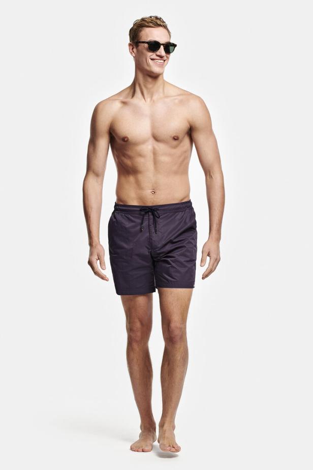 Ein Mann mit Sonnenbrille und Badehose steht vor einem weißen Hintergrund.