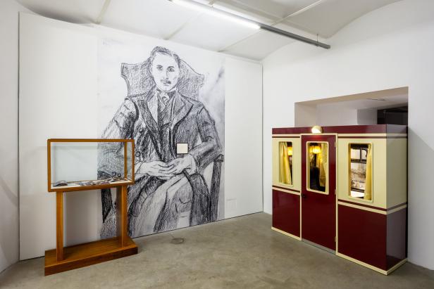 In einem Ausstellungsraum sind ein Porträt, eine Vitrine und eine Kabine zu sehen.