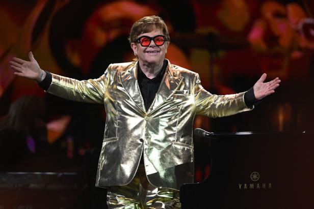Elton John in einem goldenen Anzug steht mit ausgebreiteten Armen vor einem Klavier.