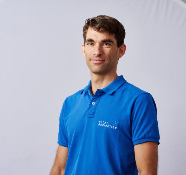 Ein Mann mit braunen Haaren trägt ein blaues Poloshirt mit dem Aufdruck „Sportordination“.