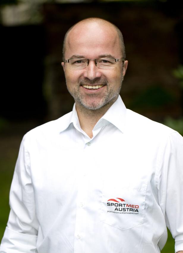 Ein lächelnder Mann mit Brille und Hemd mit dem Logo von Sportmed Austria.
