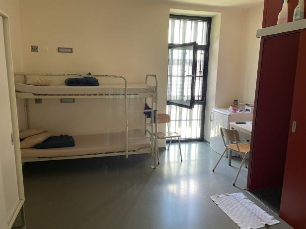 Einrichtung mit Etagenbett, Tisch und vergittertem Fenster.