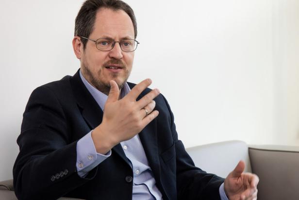 Ein Mann mit Brille im Anzug sitzt auf einem Sofa und gestikuliert beim Sprechen.