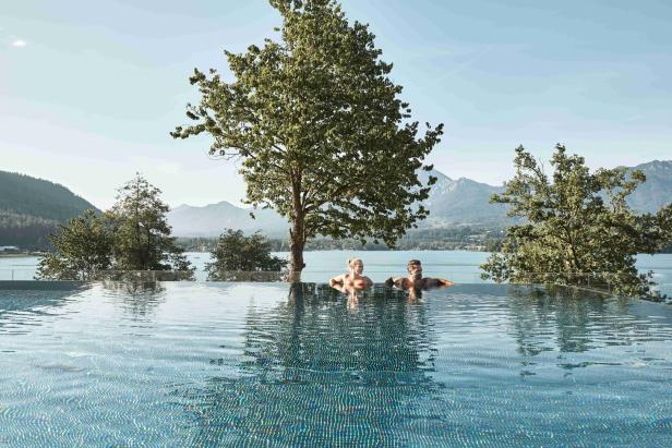 Ein Mann und Frau sitzen im Pool, hinter ihnen liegt der Faaker See und dahinter die Berge.