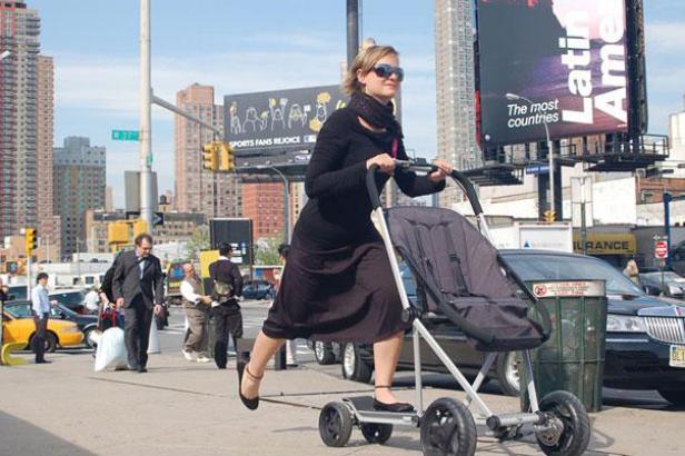 Eine Frau fährt mit einem Kinderwagen-Tretroller auf einem Bürgersteig in der Stadt.