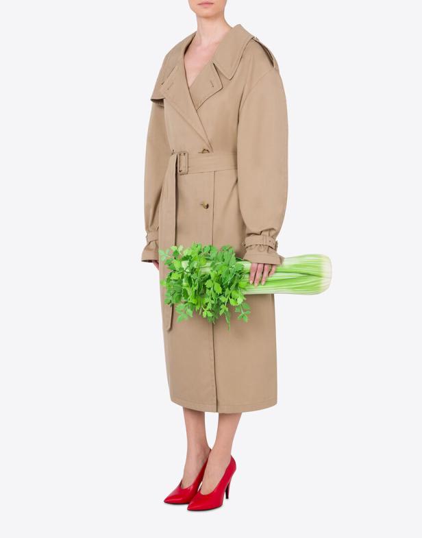 Eine Frau im Trenchcoat und roten Schuhen hält einen Bund Sellerie.