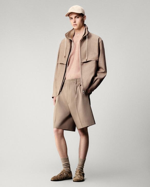 Ein Mann in beiger Jacke, Shorts, Socken und Sandalen posiert vor einem hellen Hintergrund.