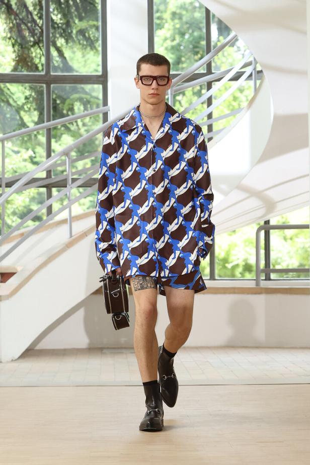 Ein Mann mit Brille, Hemd und Shorts mit Muster läuft mit einer kleinen Tasche in der Hand.