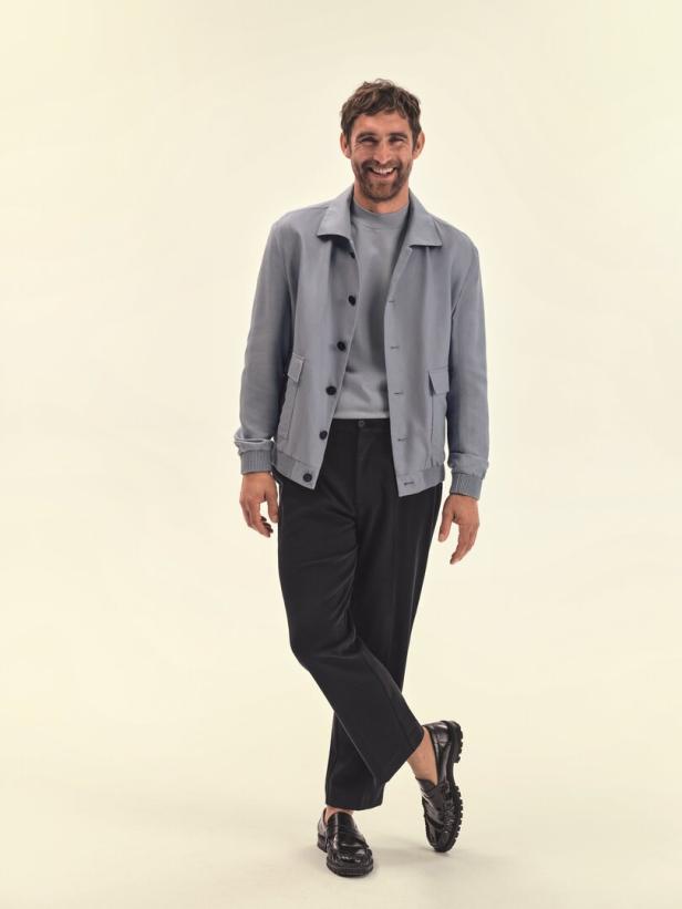 Ein lächelnder Mann in einer blauen Jacke, schwarzer Hose und schwarzen Loafers posiert vor einem hellen Hintergrund.