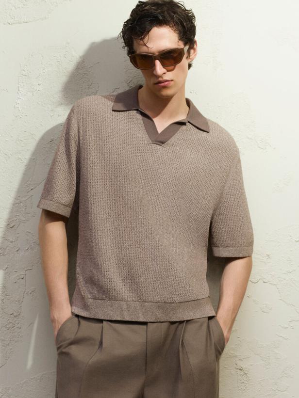 Ein Mann mit Sonnenbrille posiert in einem braunen Strickpolo und passender Hose vor einer Wand.