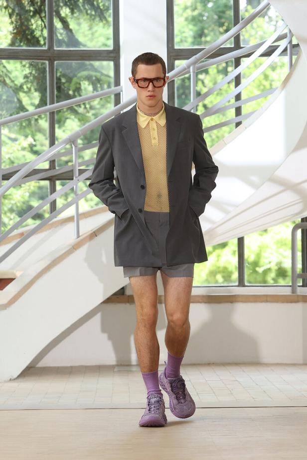 Ein Mann in Shorts, Blazer, gelbem Hemd, Socken und Turnschuhen posiert vor einer Treppe.