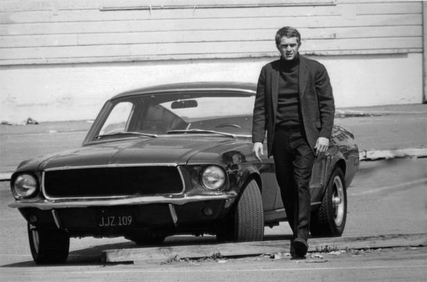 Steve McQueen steht neben einem dunklen Ford Mustang mit dem Kennzeichen JJZ 109.
