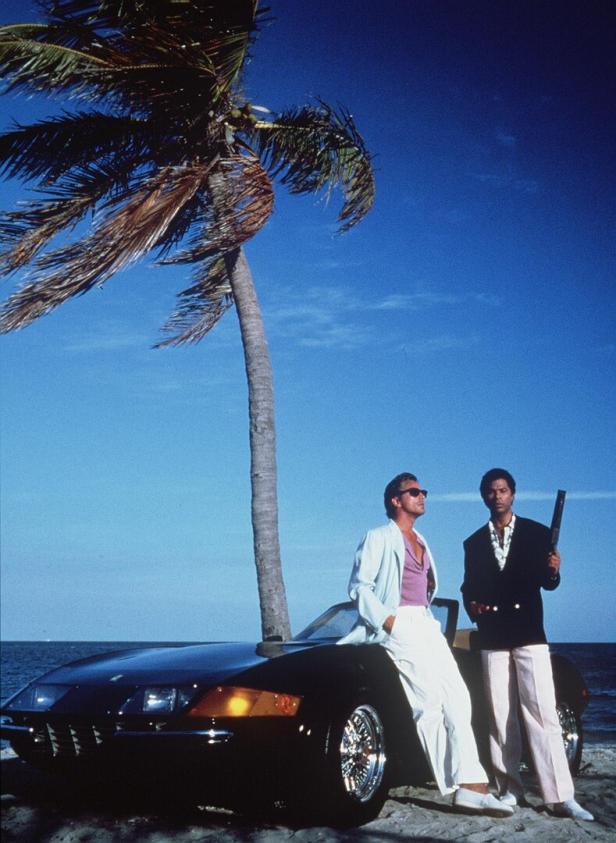 Zwei Männer im Stil der 1980er Jahre posieren mit einem Sportwagen unter einer Palme am Strand.