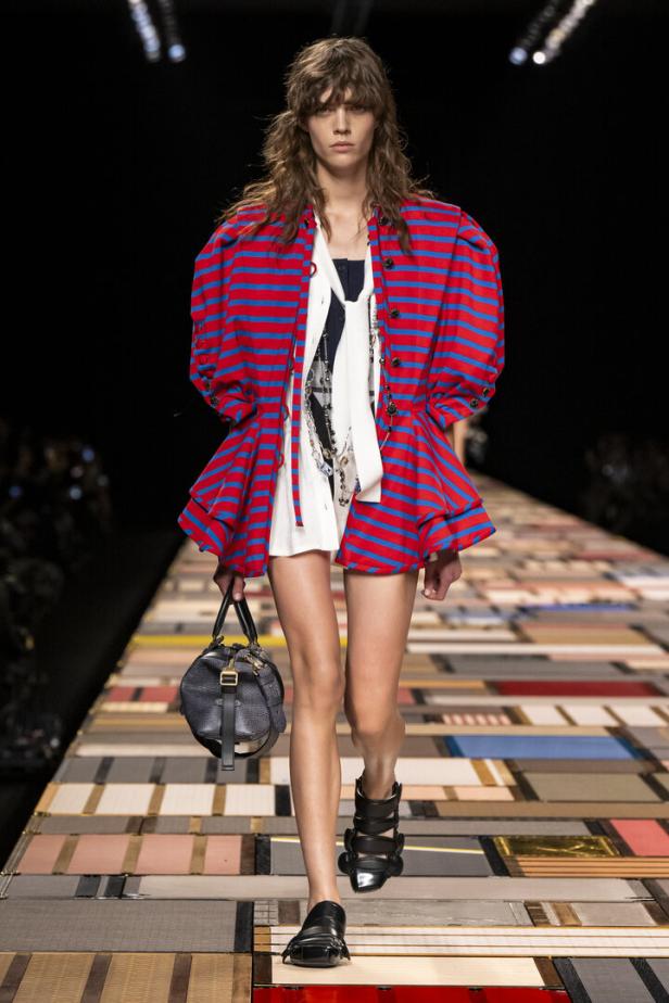 Ein Model auf dem Laufsteg mit einem rot-blau gestreiften Blazer und einer schwarzen Handtasche.