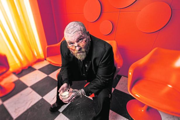 Ein tätowierter Mann mit Bart und Brille sitzt auf einem orangefarbenen Stuhl und hält ein Glas Milch.