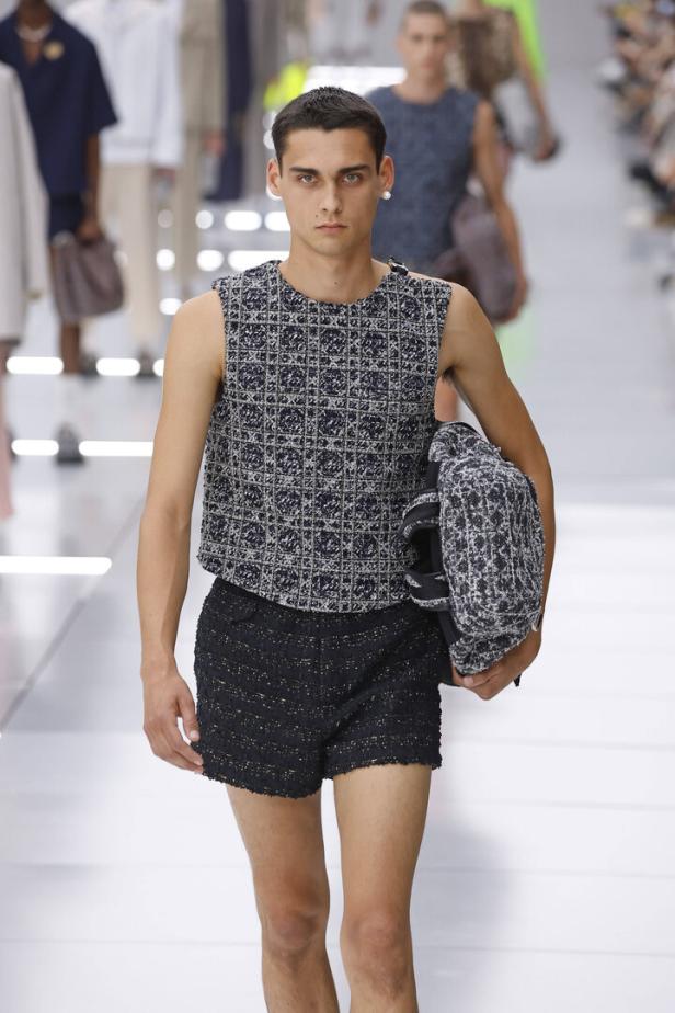 Ein Model mit gestricktem Tanktop und gestrickter Short am Laufsteg. Der junge Mann blickt ernst drein.