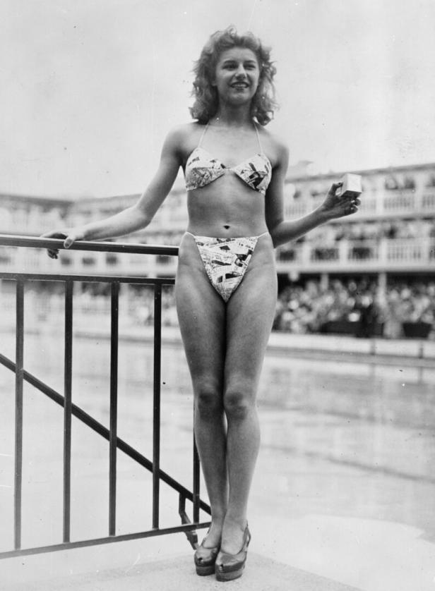 Eine junge Frau im Bikini mit Zeitungsmuster steht am Schwimmbecken.