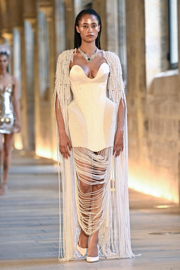 Ein Model präsentiert ein weißes Kleid mit Perlen auf dem Laufsteg einer Modenschau.
