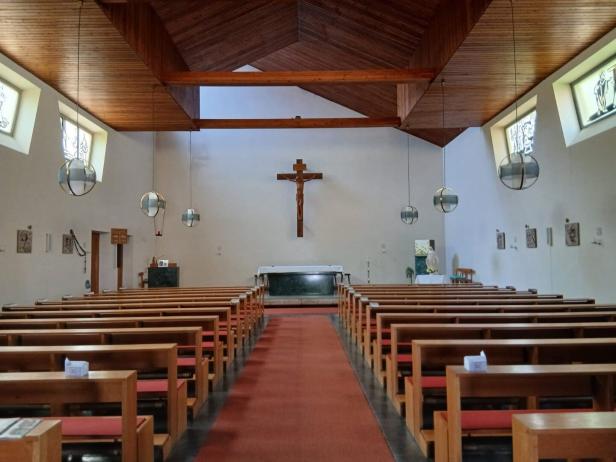 Kircheninnenraum mit Holzbänken, Mittelgang, Altar und Kreuz an der Frontwand.