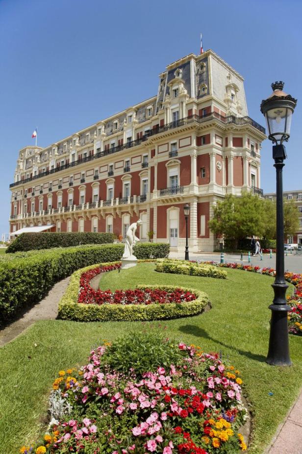 Das Hôtel du Palais in Biarritz, umgeben von einem gepflegten Garten mit Blumen und einer Statue.