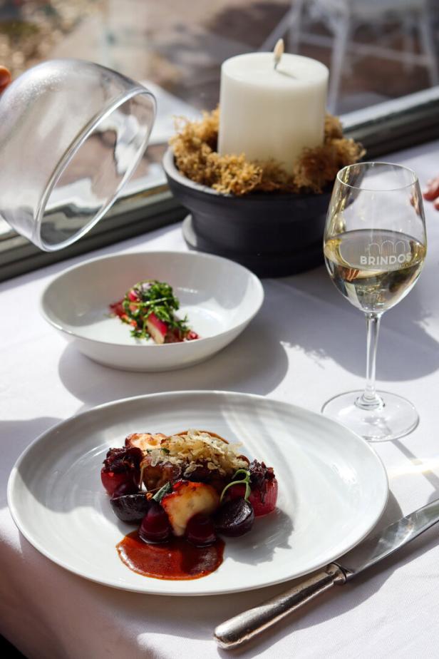 Ein Teller mit einem Gericht, bestehend aus Fleisch, Rüben und Soße, steht auf einem Tisch neben einem Glas Wein.