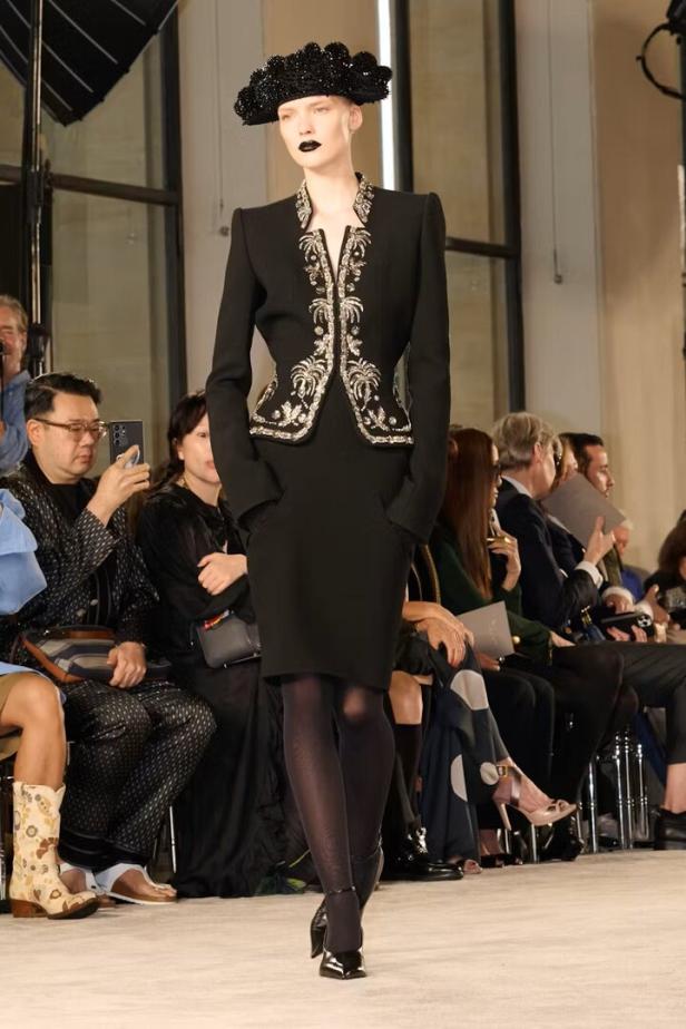 Eine Frau mit Hut und dunklem Lippenstift präsentiert ein schwarzes Kleid mit silbernen Verzierungen.