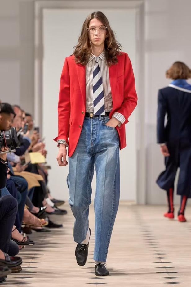 Ein Mann mit Brille, rotem Blazer und Jeans auf einem Laufsteg.