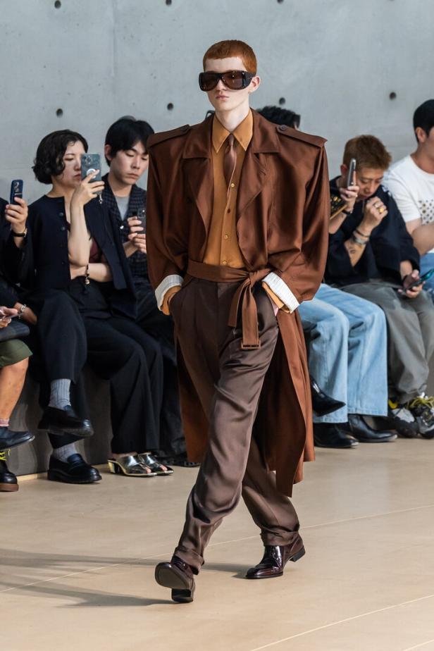 Ein Model läuft in einem braunen Trenchcoat und passender Hose über den Laufsteg von Yves Saint Laurent.