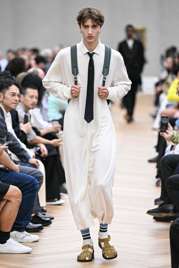 Ein Model läuft in einem weißen Overall, Sandalen und mit einem Rucksack über einen Laufsteg.