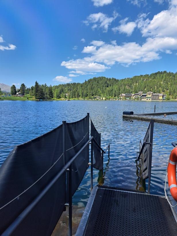 Blick von einem Steg auf einen ruhigen See mit bewaldeten Hügeln im Hintergrund.