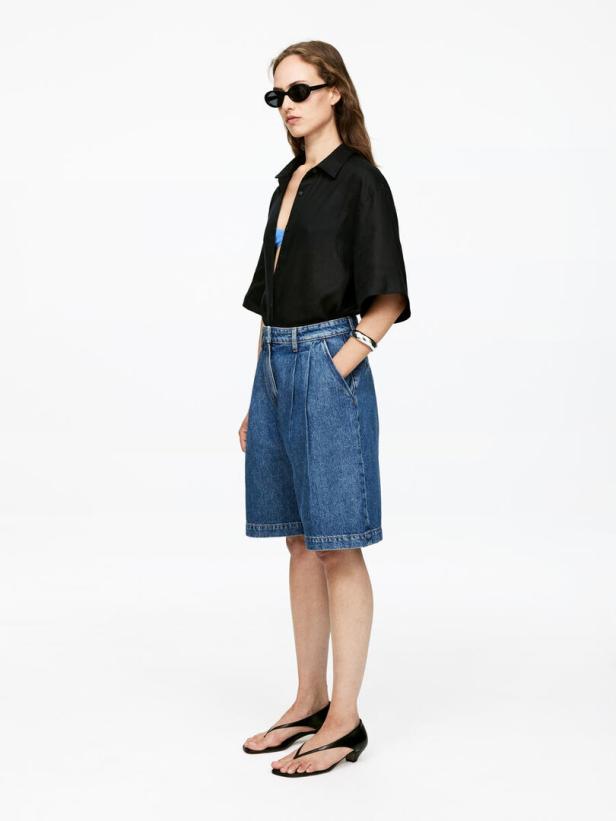 Eine Frau mit Sonnenbrille, schwarzem Hemd und Jeansshorts posiert vor einem weißen Hintergrund.