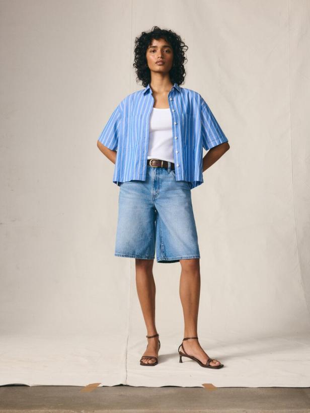 Eine Frau mit lockigem Haar trägt ein blau gestreiftes Hemd, Jeansshorts und Sandalen.