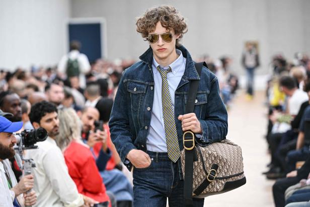 Ein Mann mit lockigem Haar trägt eine Jeansjacke und eine Dior-Tasche auf einer Modenschau.