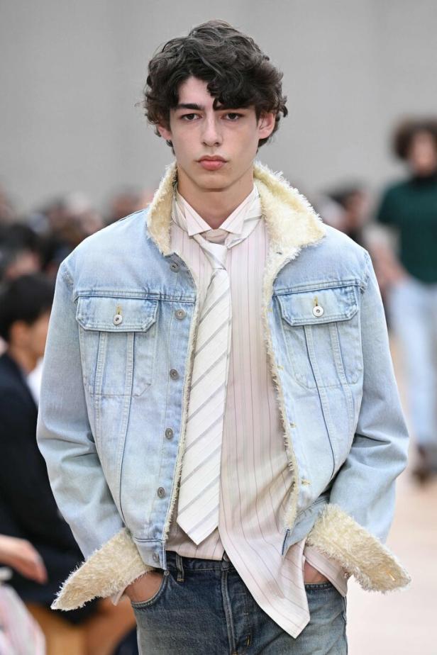 Ein junger Mann trägt eine Jeansjacke mit Fellbesatz und eine gestreifte Krawatte bei einer Dior Homme Modenschau in Frankreich.