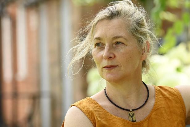 Eine Frau mit blonden Haaren und einem orangefarbenen Oberteil blickt nach links.